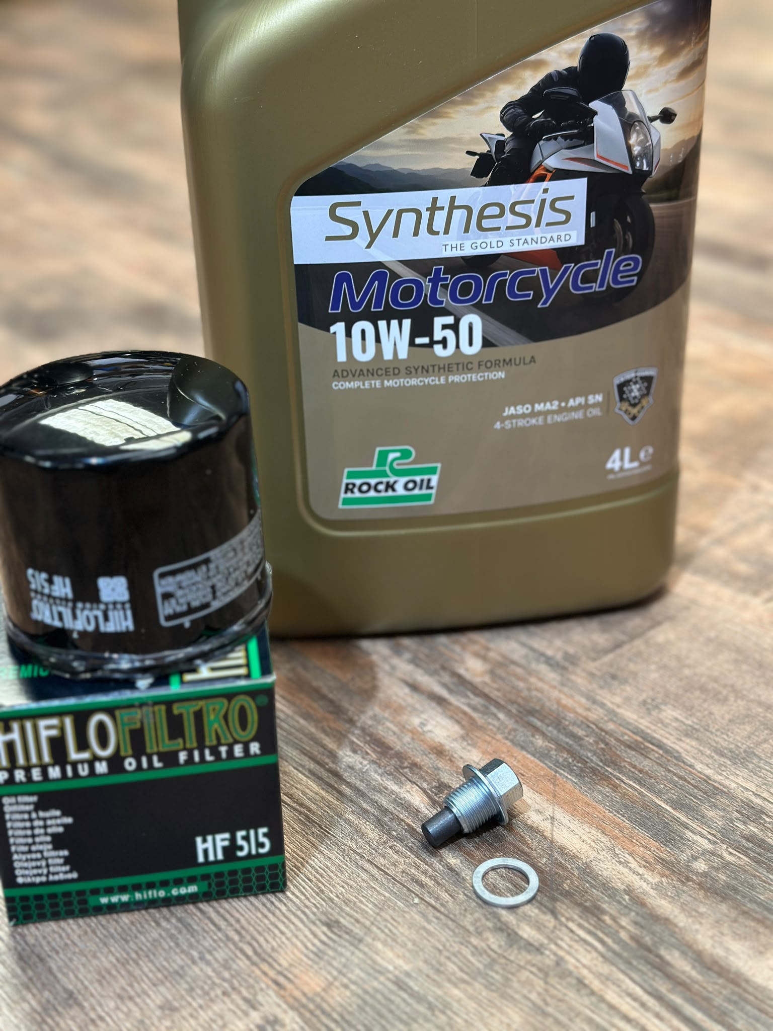 A motorcycle oil change kit on a wooden surface, including a large bottle of Rock Oil Synthesis Motorcycle 10W-50 oil, a Hiflo Filter premium oil filter in its box, and a silver bolt with a rubber gasket.