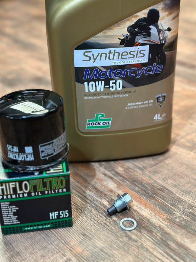 A motorcycle oil change kit on a wooden surface, including a large bottle of Rock Oil Synthesis Motorcycle 10W-50 oil, a Hiflo Filter premium oil filter in its box, and a silver bolt with a rubber gasket.